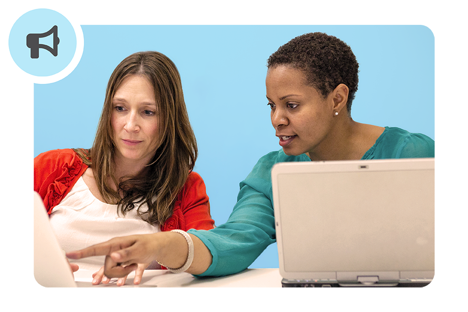 Two women sit side by side at a table, each with a laptop. One woman points at the other’s screen as they focus on the display against a plain blue background.