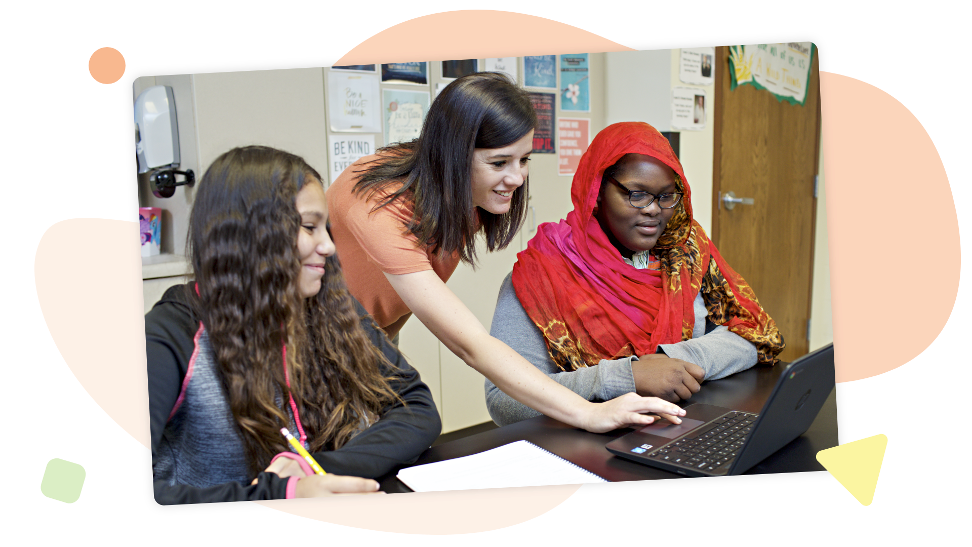 A teacher assists two students, one wearing a red headscarf, as they work together on a laptop in a classroom setting.