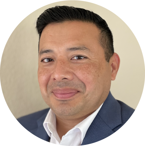 Man with short dark hair wearing a blue suit jacket and white shirt, facing the camera and smiling slightly, standing in front of a neutral background.