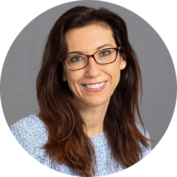 A woman with long brown hair and glasses wearing a light blue sweater, smiling at the camera against a gray background.