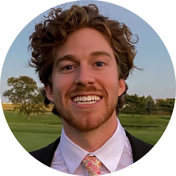 A man with curly brown hair and a beard, smiling broadly, wearing a suit, dress shirt, and floral tie, standing outdoors with trees and grass in the background.