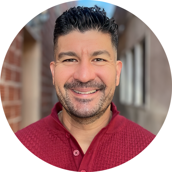 Smiling man with short dark hair and a beard, wearing a red textured sweater, standing in an outdoor setting with buildings in the background.