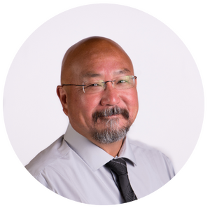A man with glasses, a shaved head, and a short beard, wearing a light gray shirt and black tie, smiles at the camera against a plain white background.