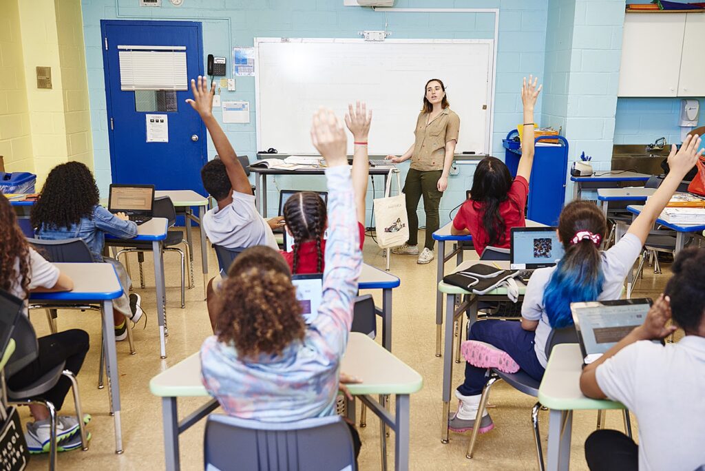 A teacher stands at the front of a classroom, while several students seated at desks raise their hands. Some students have laptops open on their desks.