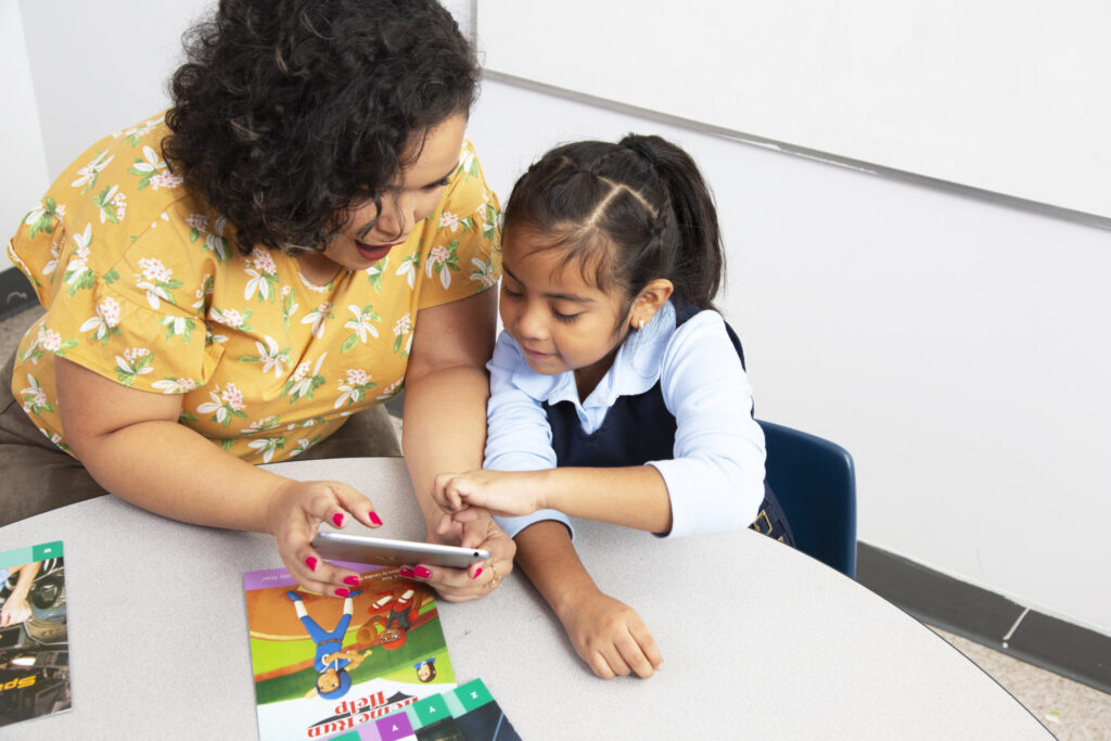 An adult and a young girl sit at a table looking at a smartphone together, with children's books on the table in front of them.