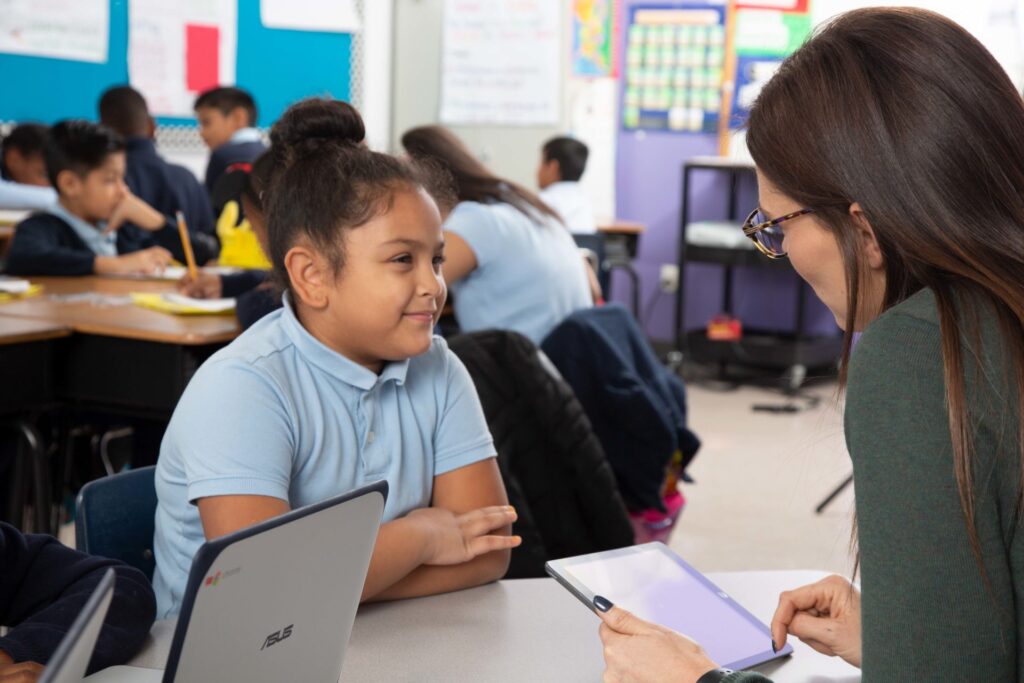 A teacher holding a tablet talks to a smiling student in a classroom. Other students are seated and working at tables in the background.