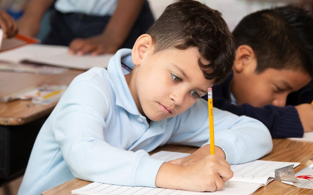 A young boy in a light blue shirt writes in a notebook at his desk in a classroom, with other students working nearby.