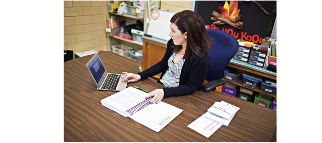 A woman sits at a desk in a classroom, looking at a laptop while referencing an open binder and papers.