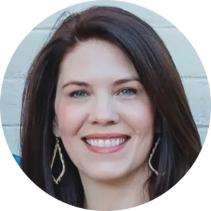 Woman with long brown hair and light skin smiling, wearing gold earrings, in front of a light-colored brick wall.