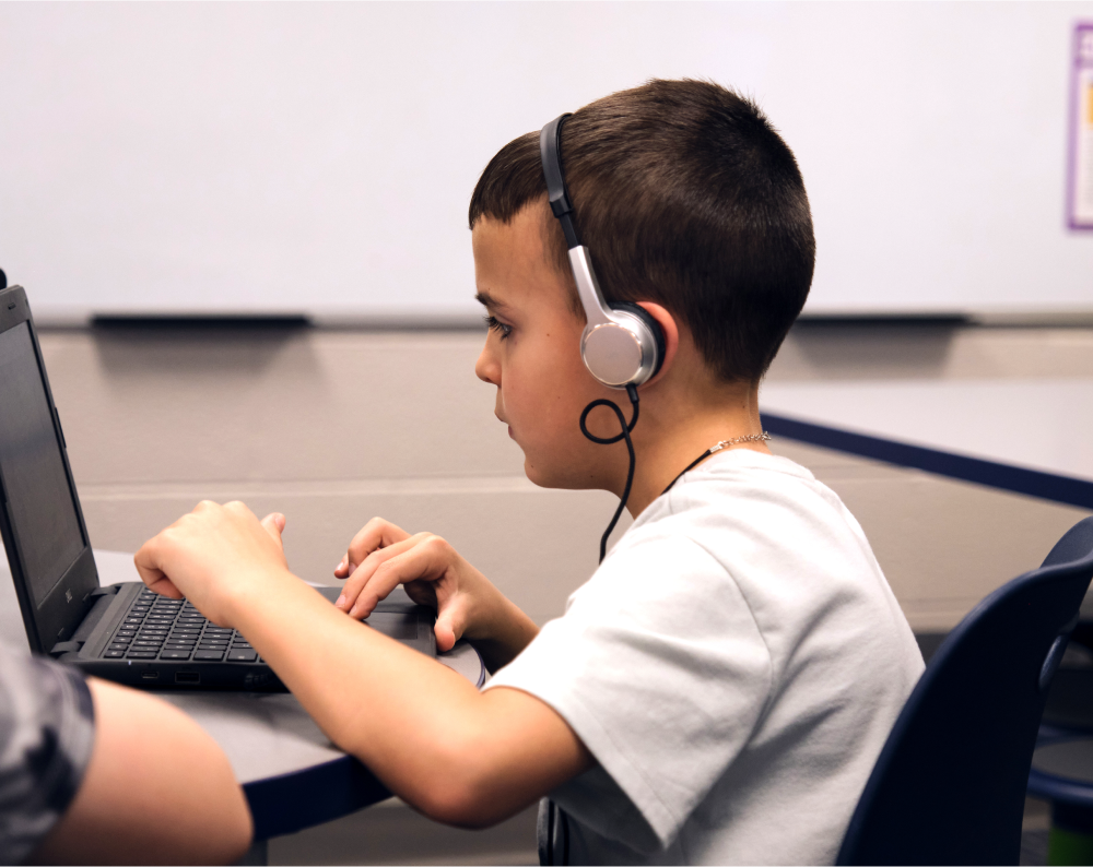 A young boy wearing headphones uses a laptop computer at a desk in a classroom, exploring Amplify programs that are grounded in evidence based programs and research and case studies.