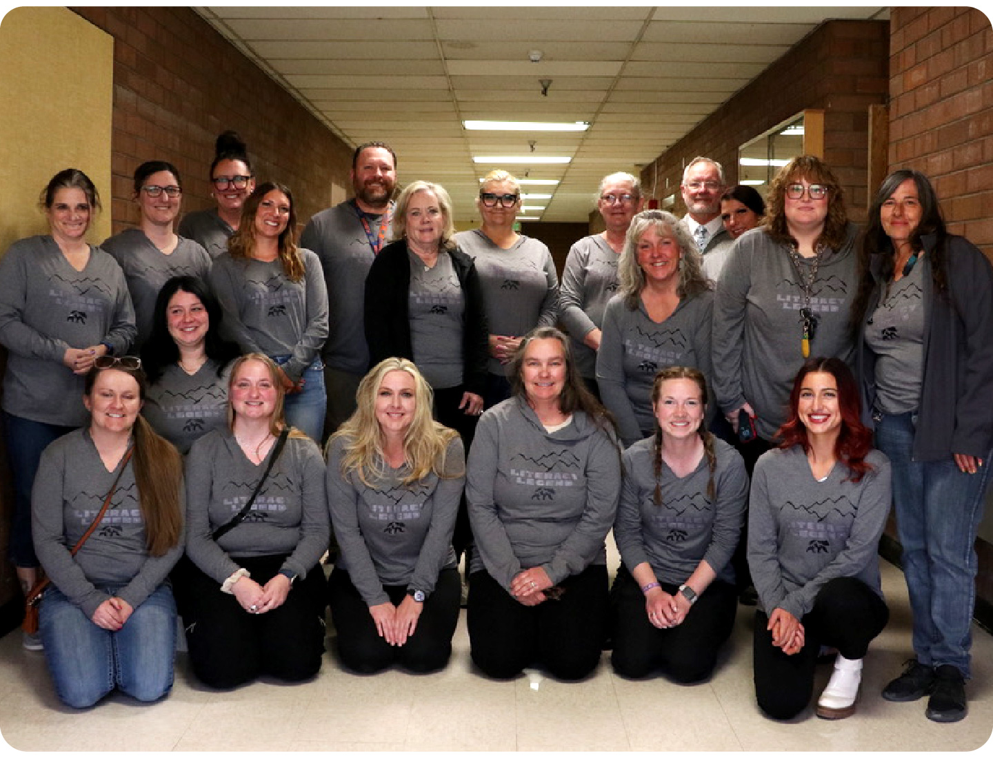 A group of adults poses in a hallway, all wearing matching gray shirts with a black design, celebrating their work with early literacy and the Science of Reading curriculum. Most are smiling and kneeling or standing in two rows.