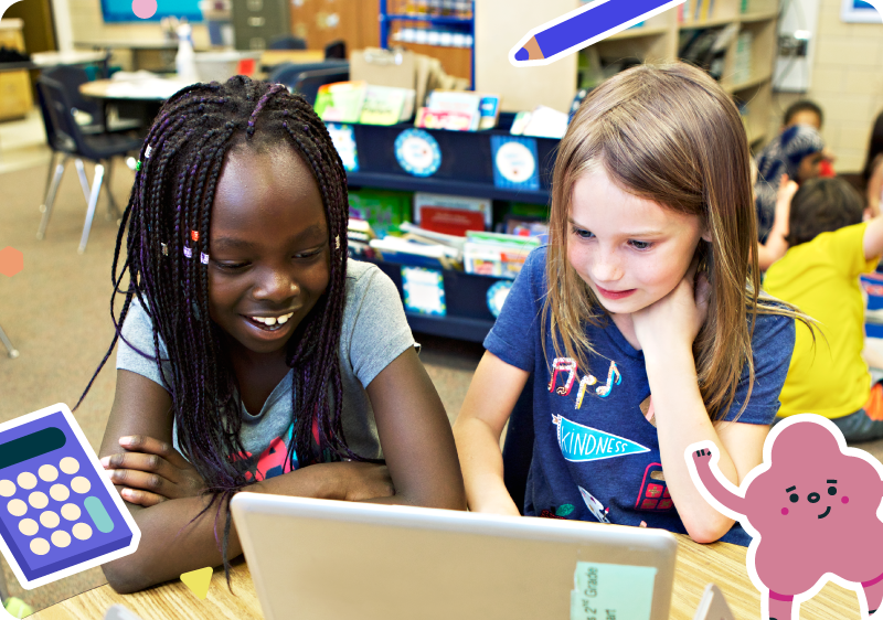 Two elementary school girls sit together at a classroom table, looking at a laptop screen and smiling, with bookshelves and other students in the background.