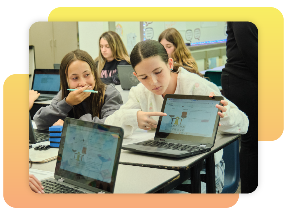 Two students sit at desks using laptops in a classroom, one pointing at her screen while the other looks on; more students are visible in the background.